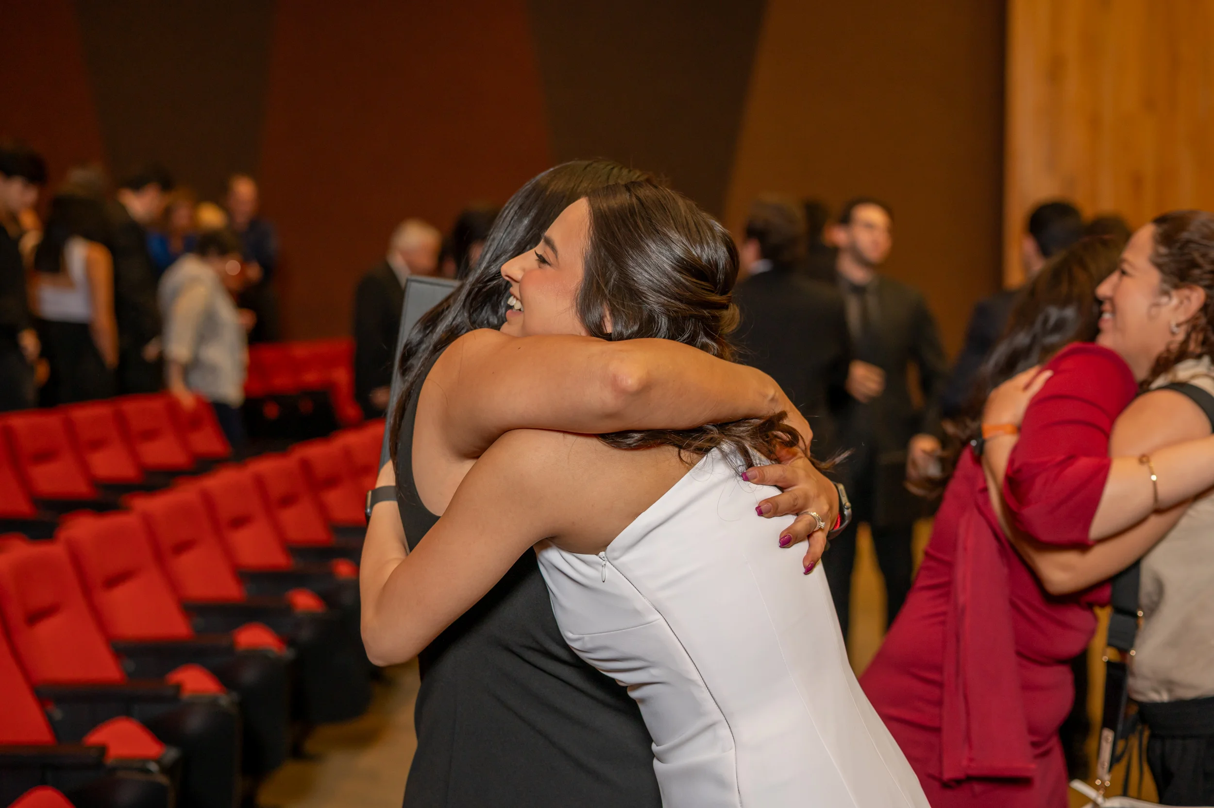 Dos egresadas con vestido de gala se abrazan sonriendo en el auditorio de la FLDM tras la ceremonia de graduacion, con otro abrazo visible al fondo